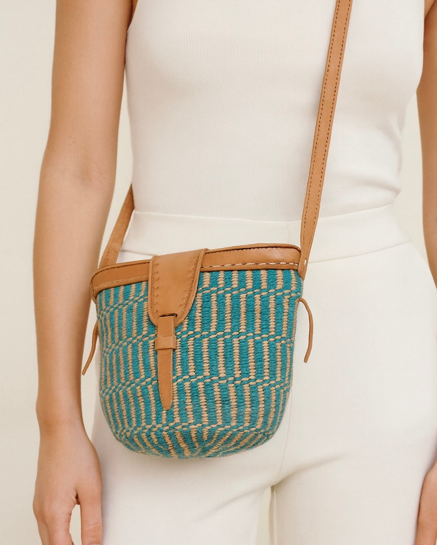 Person wearing a teal woven crossbody bag with brown leather strap on a plain background